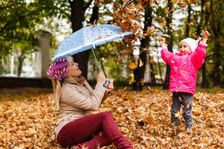 快乐的妇女拿着雨伞, 和她的小女儿玩秋季树叶在城市公园的乐趣