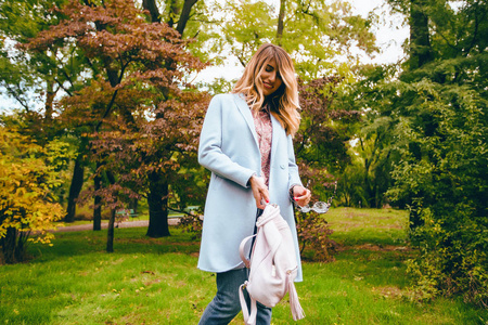 穿着蓝色大衣的快乐年轻女子的户外肖像在秋季公园举行时尚粉红色背包摆姿势