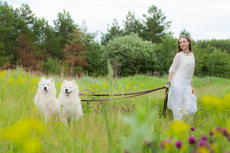 年轻的美丽的女人站在绿色的草地上的森林背景上的两个白色毛茸茸的大狗
