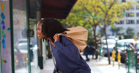 漂亮的黑人女性拿着购物袋看着商店窗口微笑