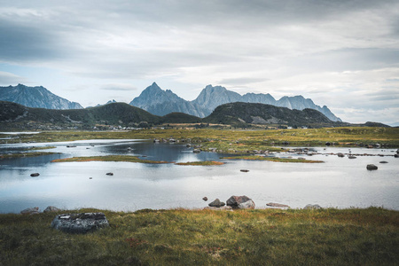 山在清澈的水面上的神奇的倒影在多云天。水与草在前景, 山在背景。在挪威北部的夏季旅行。罗弗敦群岛