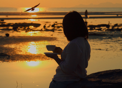 剪影女人拿着咖啡看着海鸥飞在海景日落