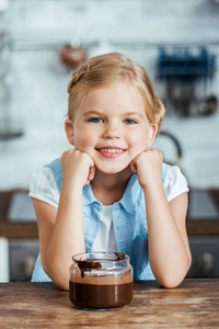 可爱的快乐的孩子坐在桌子与巧克力在罐子里传播和微笑在照相机