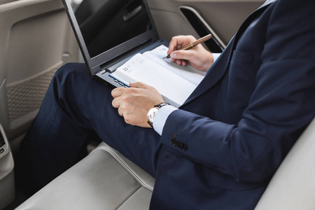 一个穿着西装的年轻商人坐在一辆商务车的后座上, 拿着笔记本电脑和笔记本, 进行商务谈判