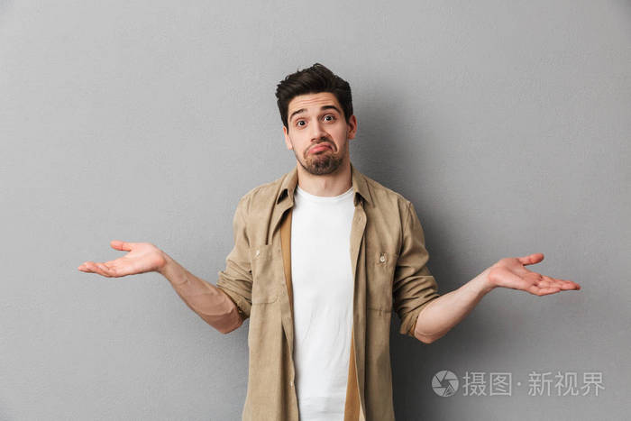 一个困惑的年轻的不经意耸耸肩肩膀孤立在灰色背景的肖像