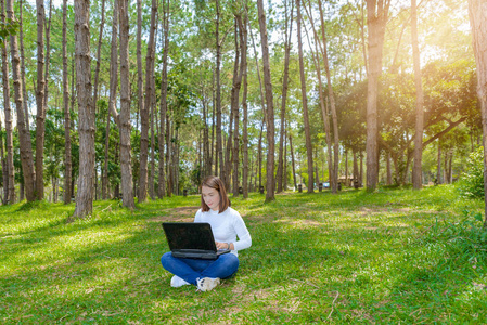 美丽的年轻女子的肖像坐在绿色草地在公园与腿交叉在夏季白天, 而使用笔记本电脑。背景是松树林的景观