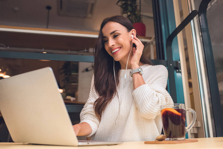 微笑的年轻女性自由职业者的耳机工作在笔记本电脑在餐桌上, 在咖啡馆的品酒