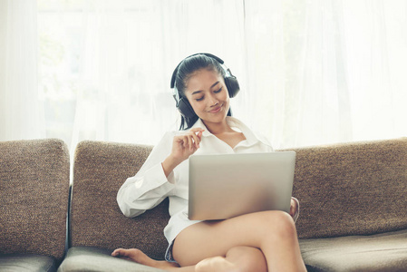 穿着白衬衫的开朗的年轻女子坐在沙发上听音乐从耳机, 而在家里手持笔记本电脑