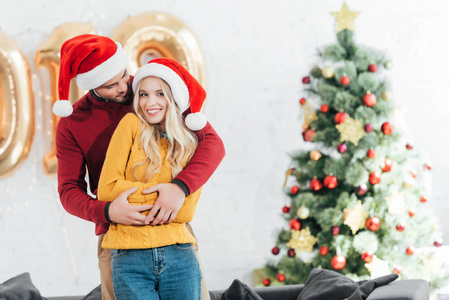 快乐的人拥抱他的微笑的女朋友在家与圣诞树
