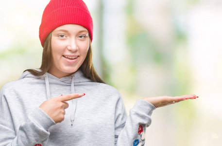 年轻的高加索美丽的女人穿着羊毛帽在孤立的背景惊讶和微笑的相机, 同时提出用手和手指指指点点