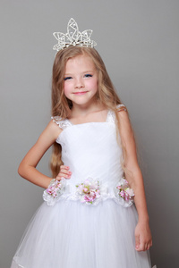 戴着一顶王冠和白的可爱小女孩打扮可爱的微笑和冒充为灰色的背景上的摄像头
