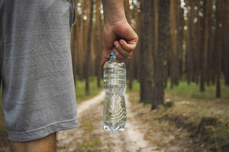 人拿着一瓶水在泥土的道路在树林的背景。在森林和山脉的户外和远足的训练概念
