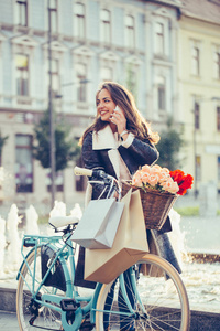 美丽的年轻微笑的妇女与购物袋, 倚在她的自行车和说话的手机