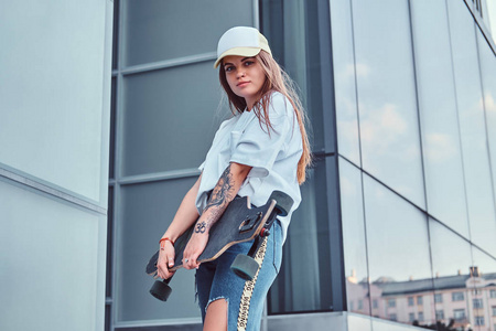 年轻的时髦女孩在帽子身着白色衬衫和撕裂牛仔裤举行滑板, 而在摩天大楼附近的姿势