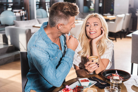 美丽的金发女郎拿着杯咖啡, 在咖啡馆里看着男朋友。