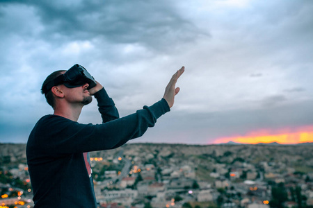 一个人在虚拟现实眼镜背景上的日落在城市上空。未来技术的概念。现代技术