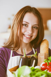 年轻女子捧着与蔬菜站在厨房里的食品杂货店购物袋