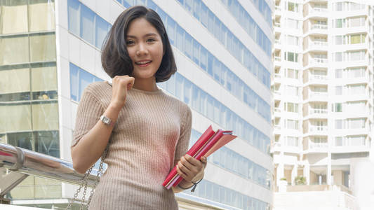 亚洲少女肖像在户外公共空间与现代建筑背景下举行红色书立场和礼服在街道时尚布料