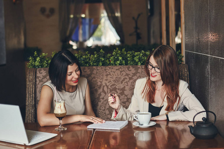 两个年轻的微笑与笔记本电脑的白种人女企业家。在咖啡馆中的妇女