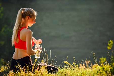 在红色的顶部和黑色裤子的年轻金发女郎坐在自然的草地上。一个运动的女人拿着一瓶水