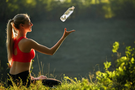 在红顶的年轻金发女郎坐在自然的草地上。一个运动的女人在她手中抛出一瓶水