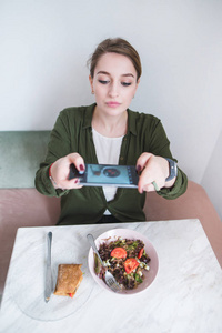 一个年轻漂亮的女人在餐厅拍了一张她的食物照片。一位博客作者在智能手机上拍摄三明治和沙拉。