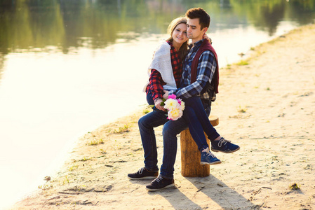 穿着衬衫和牛仔裤时尚年轻夫妇坐在湖岸上树桩上。美丽迷人的女孩坐在膝盖的帅气男友