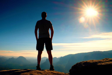 黑色的裤子的独自徒步旅行者站在岩石帝国和雾有雾的早晨谷到太阳，注视