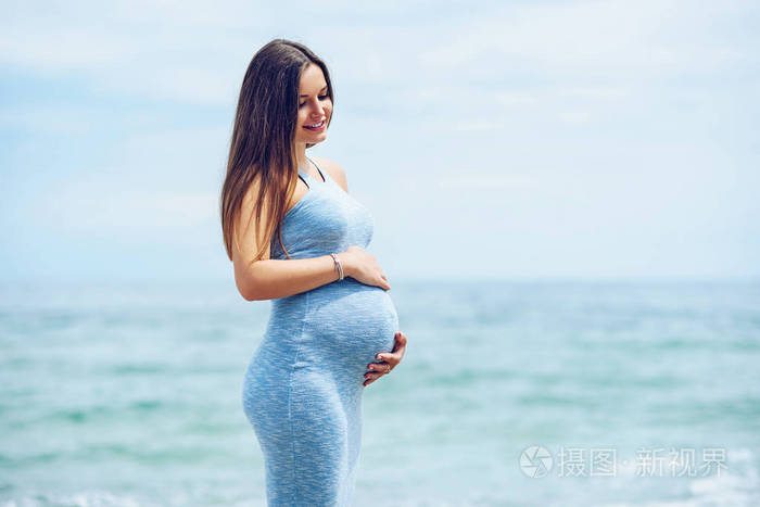 一个美丽的怀孕女人穿着蓝色紧身连衣裙上海滩摸她的肚子爱与关怀.