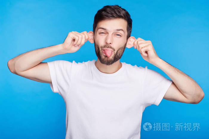 有趣的年轻白种人鬼脸扮鬼脸,伸着舌头在相机,使耳朵伸出在试图让他哭