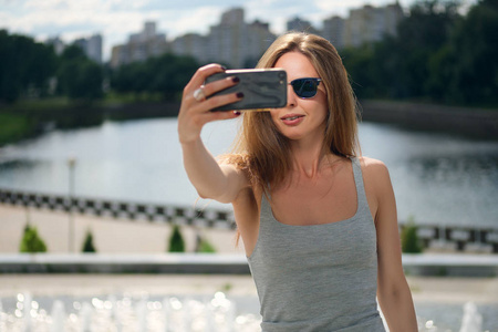 有吸引力的旅游女孩与河岸上背景的自拍照在城市公园