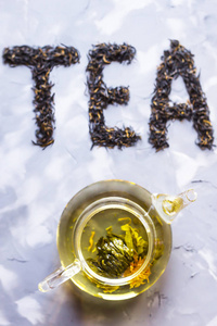 透明茶壶与绿茶旁边的题词 茶 奠定了干燥的茶叶在灰色的具体背景