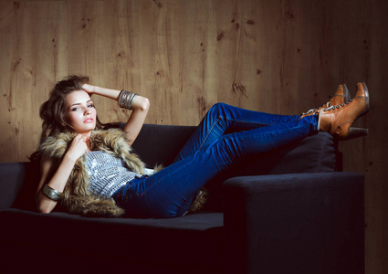 优雅的女人坐在黑色的沙发上，穿着一件蓝色牛仔裤的肖像和毛皮背心