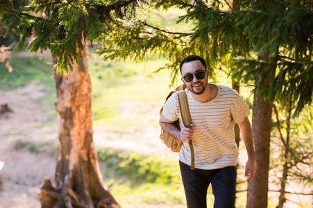 在树林里旅行英俊时尚的年轻人，穿着白色 t 恤及太阳眼镜，背包里的在他的手。在附近树上的相机摆姿势