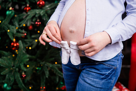 一个怀孕的女人拥有她手儿童袜子，赃物背景下的一棵圣诞树。身穿一件衬衫和帆船。特写
