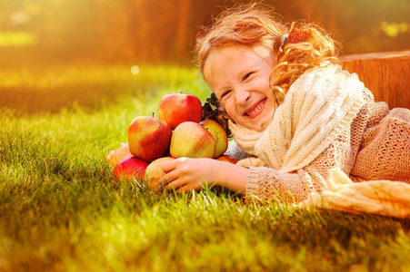 快乐的孩子女孩在白色针织围巾与红苹果在阳光明媚的秋日花园