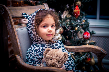 漂亮的小女孩微笑着靠近圣诞树坐在老式椅子上的玩具熊。新年快乐