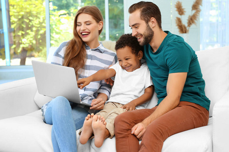 幸福的夫妇同坐在沙发上在家里上使用笔记本电脑通过非洲裔美国男孩