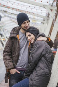 美丽的白种女人与一个男人在欧洲城市的冬天散步。夫妇穿着休闲灰色的衣服和拥抱对方。寒冷的天气, 背景下的圣诞灯