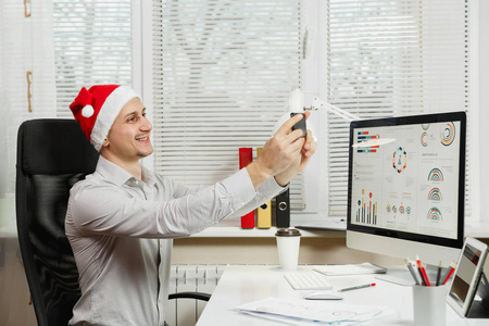 英俊的微笑商业男子在衬衫和红色圣诞帽坐在办公桌上工作地点, 做自拍在手机上, 在计算机与现代显示器工作, 在轻型办公室的文件