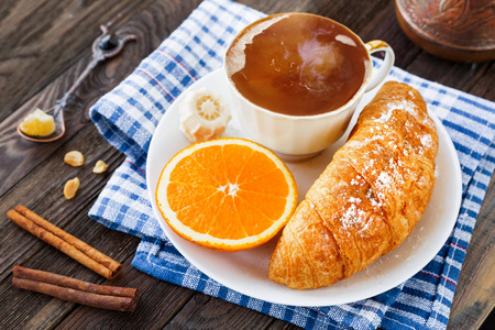 欧式早餐杯热咖啡，牛角包和橙色。美味的食物上格子蓝色餐巾，仿古木制背景