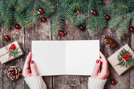 写一封信给与圣诞礼物，树皮纹理，冷杉的枝条，松果，红色装饰的木制背景的女性手。圣诞节和新年快乐卡。平躺，顶视图