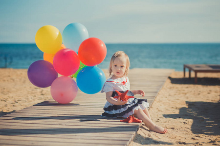 可爱的金发碧眼的女婴摆在沙滩海边的沙滩上, 在木码头上, 五颜六色的气球穿着休闲服装赤脚