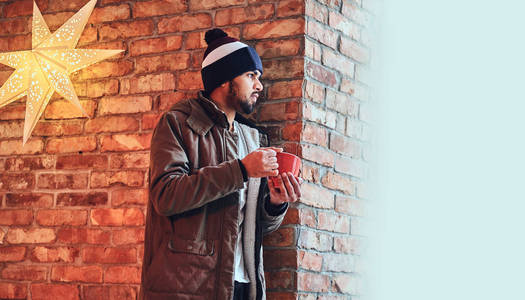 印有胡子的印度时髦男人的肖像穿着暖和的夹克和帽子, 从红砖墙边的红杯子里喝咖啡