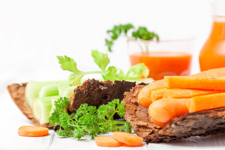 胡萝卜汁在美丽的眼镜, 切胡萝卜和芹菜在木树皮碗和绿色欧芹的白色木质背景。新鲜蔬菜饮料。关闭摄影。选择性焦点水平横幅