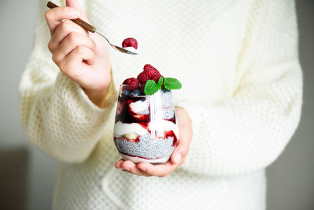 穿着白色毛衣的女孩拿着玻璃和嘉奶甜点, 覆盆子, 蓝莓, 薄荷。健康的早餐概念。复制空间, 关闭