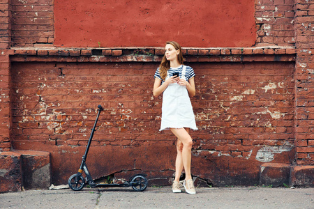 年轻漂亮的女孩站在一个红色的砖墙在夏天在城市和正在谈论的电话旁边的一辆摩托车。健康生活方式的概念