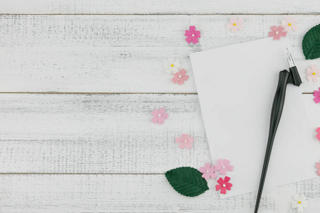 空白白色卡片和倾斜的笔装饰与粉红色纸花和绿叶在白色木头背景