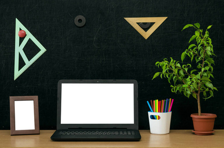 老师或学生桌。教育背景。教育理念。带空白屏幕的笔记本电脑, 绿色植物树, 书 抄写, 彩色铅笔, 黑板上的相框