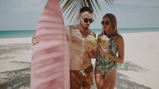 年轻快乐情侣泳装与粉红色冲浪板喝椰奶, 享受阳光明媚的夏日在他们的海滩热带假日